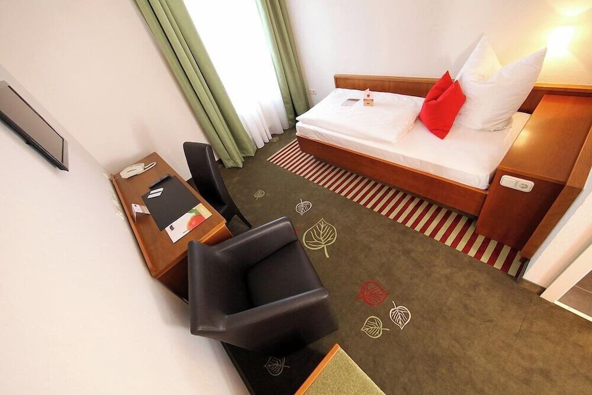 A hotel room with a bed, a desk with a telephone, a chair, and a carpet with leaf patterns.