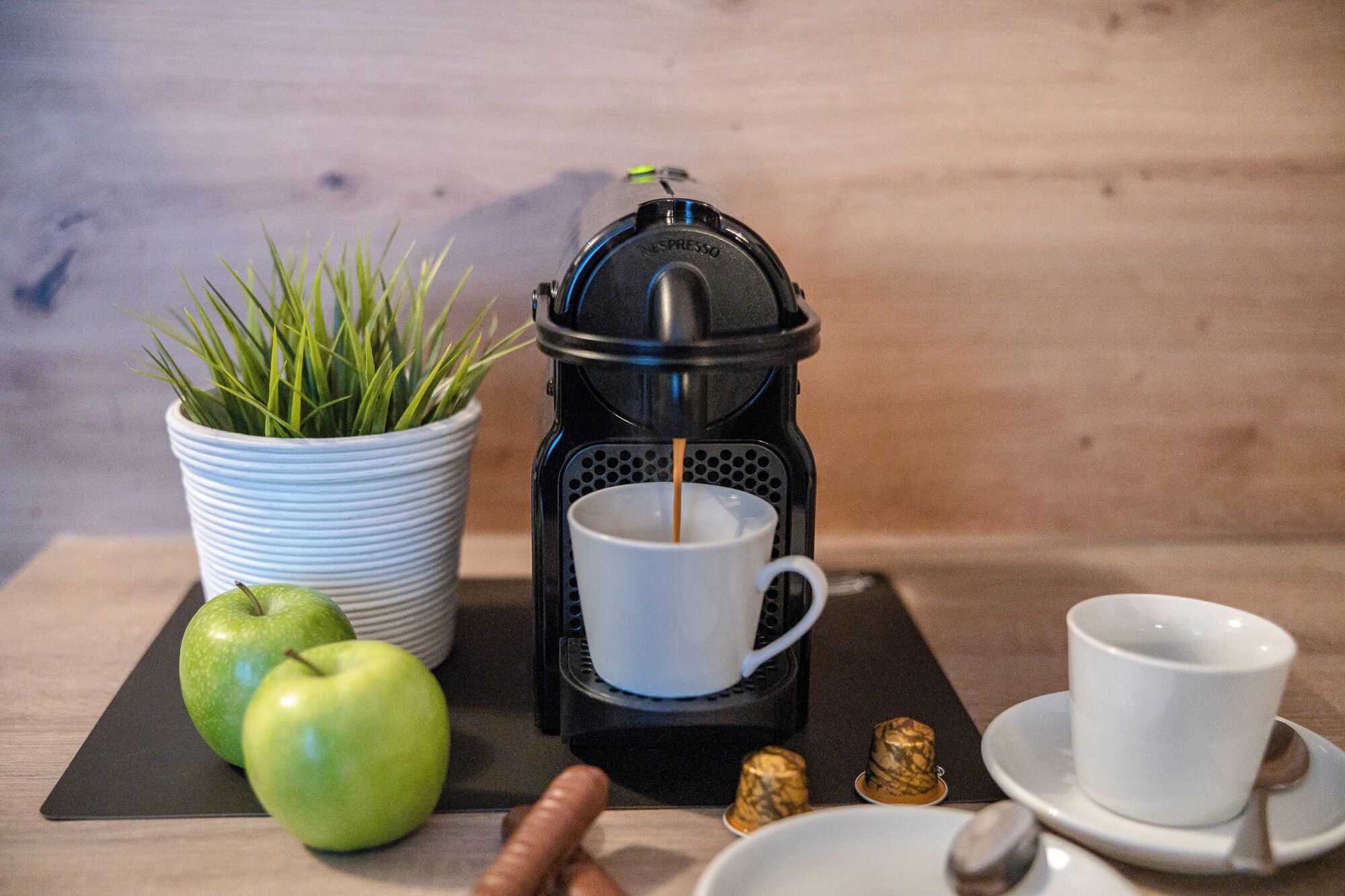 Una cafetera, una taza, un plato con una cuchara, una planta pequeña y dos manzanas sobre una mesa.