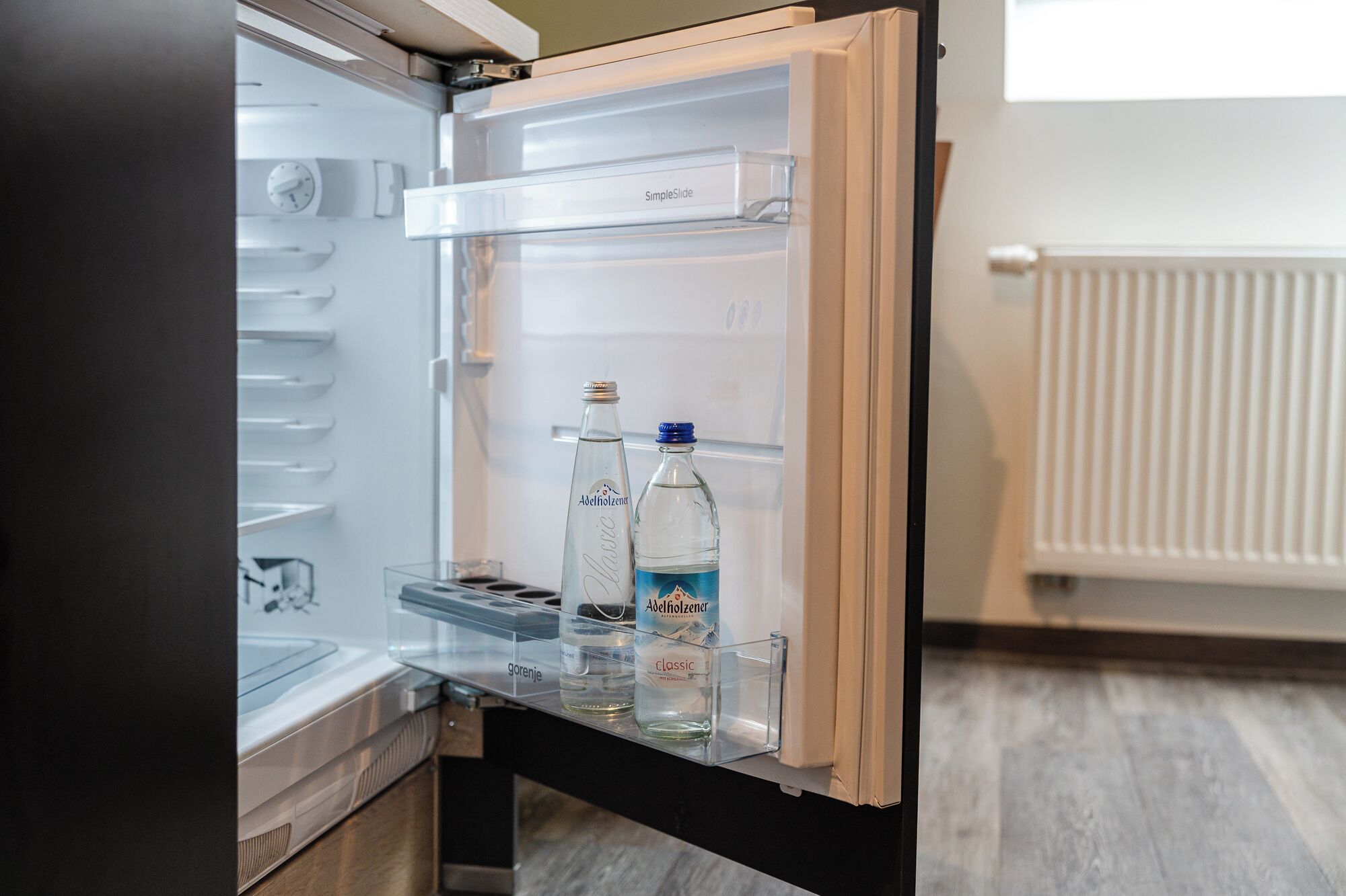 An open refrigerator with a water dispenser and two bottles labeled 'Adelholzener'.