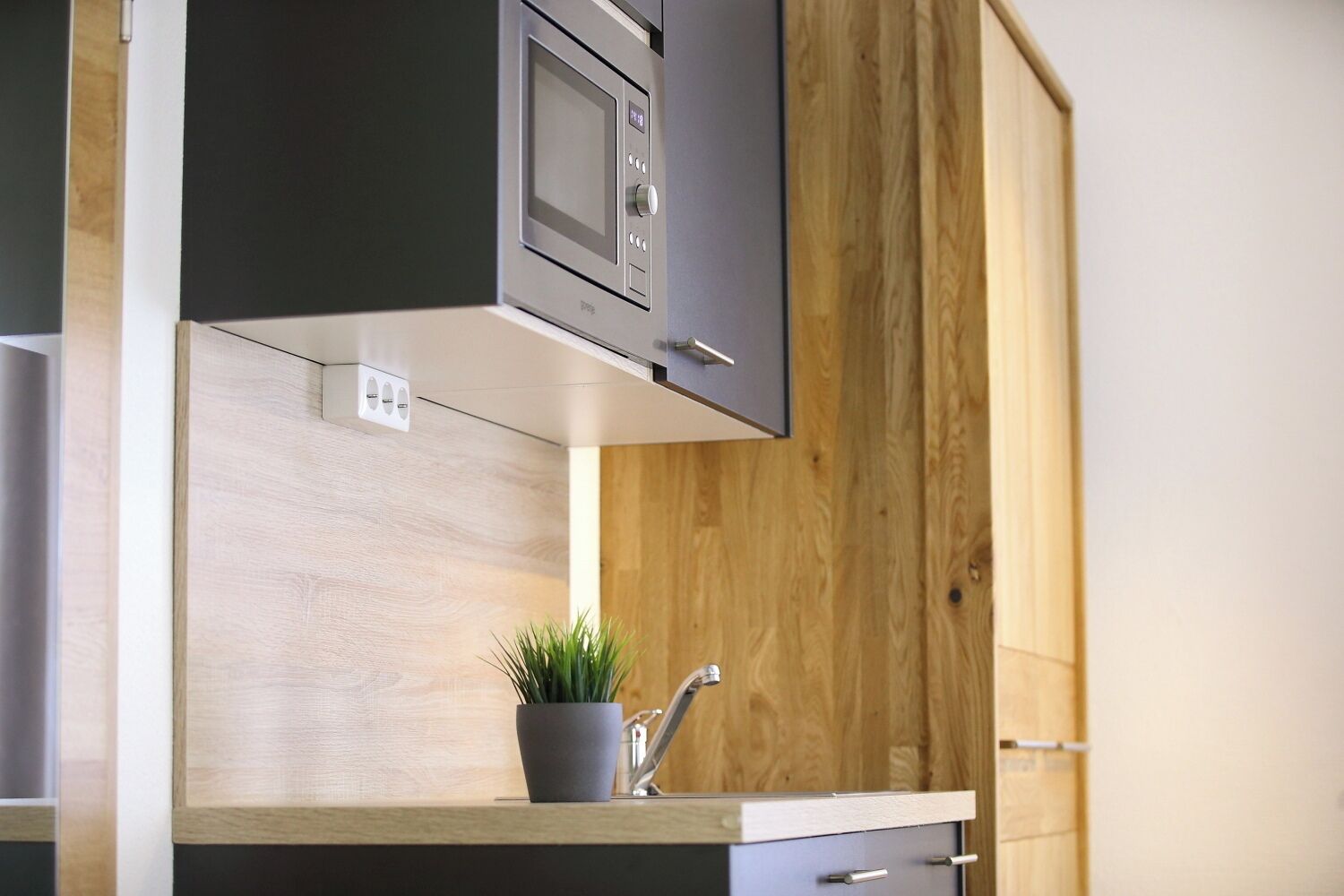 A modern kitchen with a built-in microwave, a wooden cabinet, a potted plant, and a stainless steel faucet.