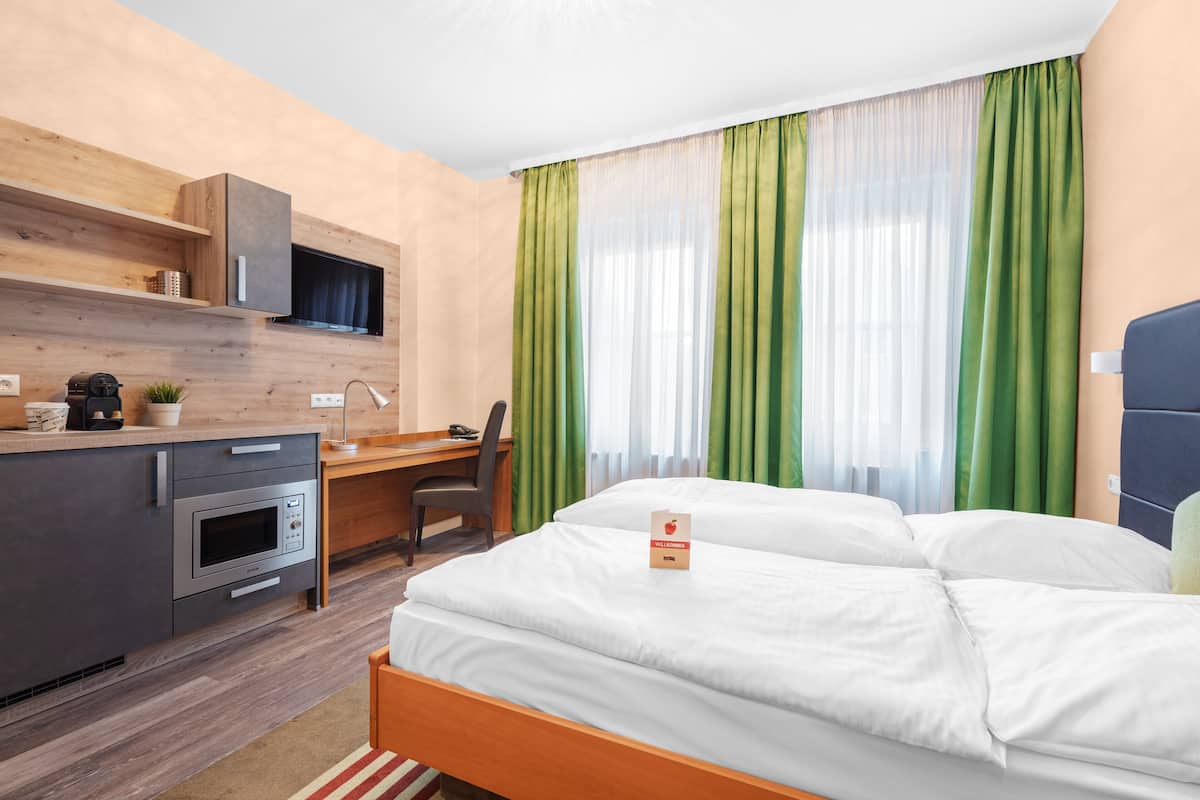A hotel room with a bed, a desk with a chair, a kitchenette, and a window with green curtains.