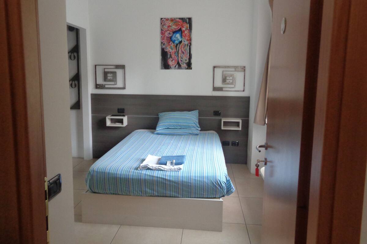 Un dormitorio con cama, mesitas de noche, un cuadro y una puerta.