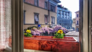 Un balcón con macetas y flores, vistas a edificios, y una persona con una chaqueta azul.