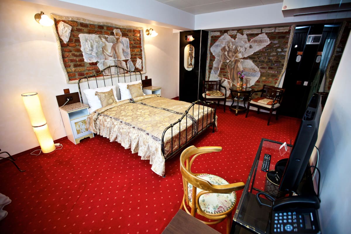 A hotel room with a red carpet, a bed with a metal frame, a wooden chair, a desk with a telephone, and a mirror on the wall.