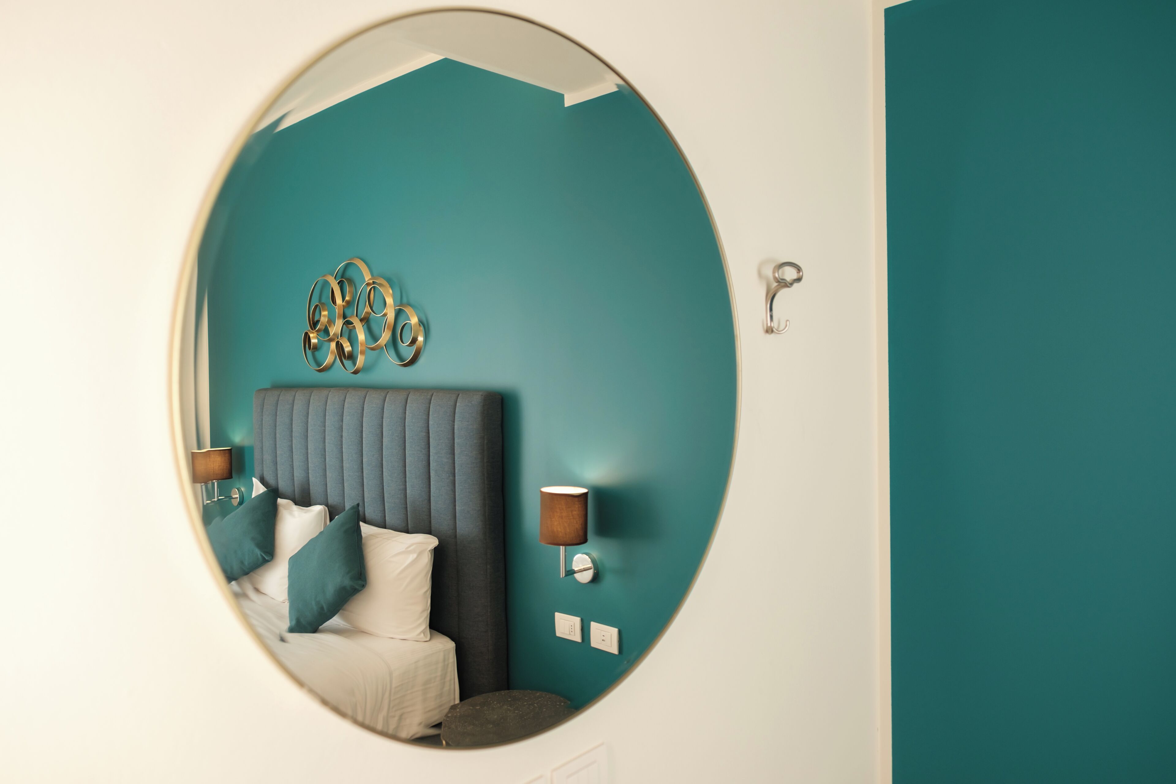 Een ronde spiegel die een bed met een gestuufd hoofdbord, een wandlamp en een decoratief wandobject weerspiegelt.