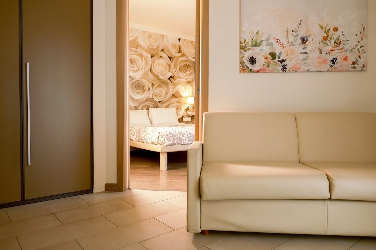Une pièce avec un canapé beige, un lit orné d’un papier peint à motifs floraux et une porte donnant sur une autre pièce.