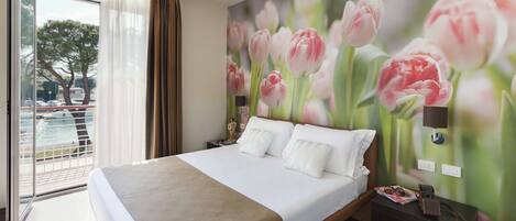A hotel room with a large bed, a nightstand with a lamp, a window with curtains, and a wall mural of flowers.