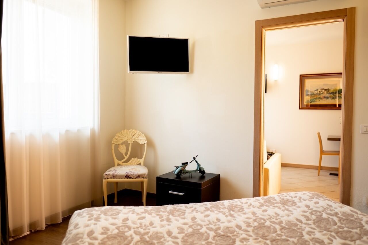 A bedroom with a bed, a chair, a small table with a scooter, a TV on the wall, and a door leading to another room.