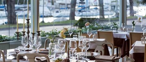 Comedor con mesas dispuestas para comer, sillas y vistas a barcos a través de amplias ventanas.
