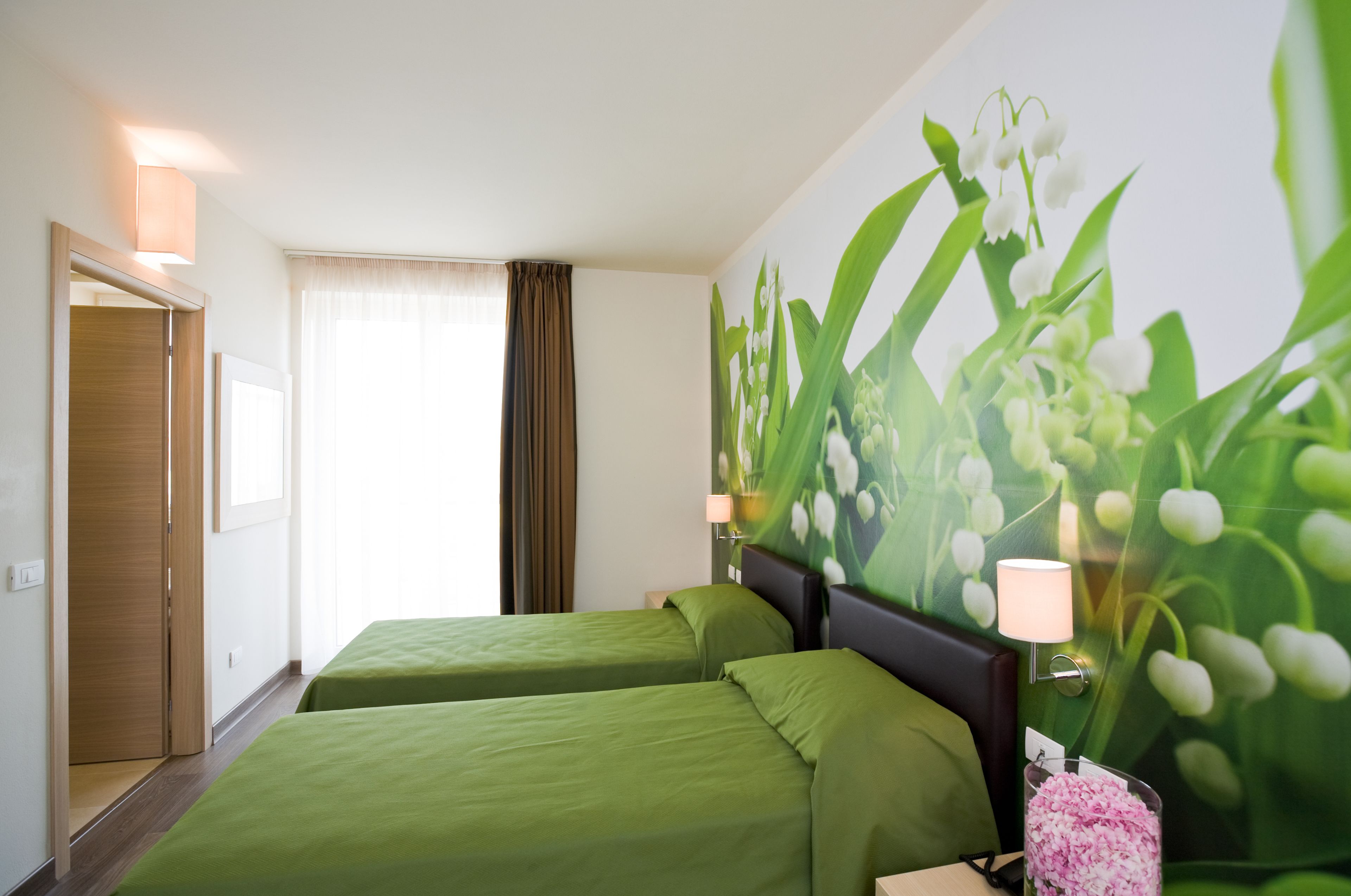 A hotel room with two beds, a large window with curtains, a wall-mounted lamp, and a floral mural.