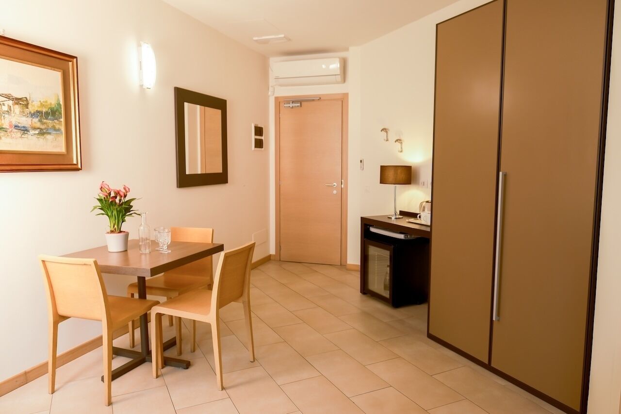 Ein Zimmer mit Esstisch, Stühlen, einer Topfpflanze, einem Spiegel, einem Kleiderschrank, einer kleinen Kommode und einer Tür.