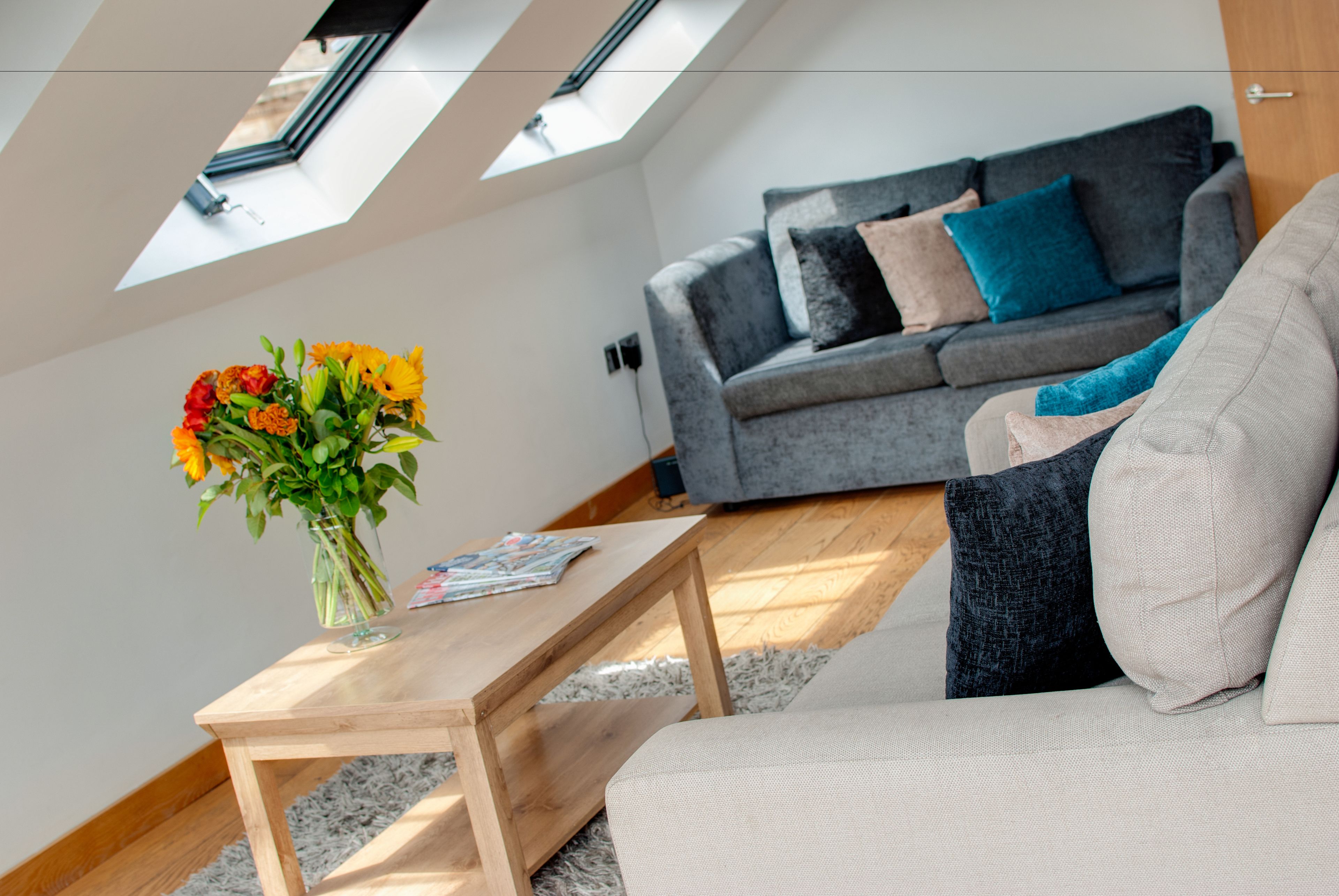 A modern living room with a grey sofa, a wooden coffee table with a vase of flowers, and a skylight above.