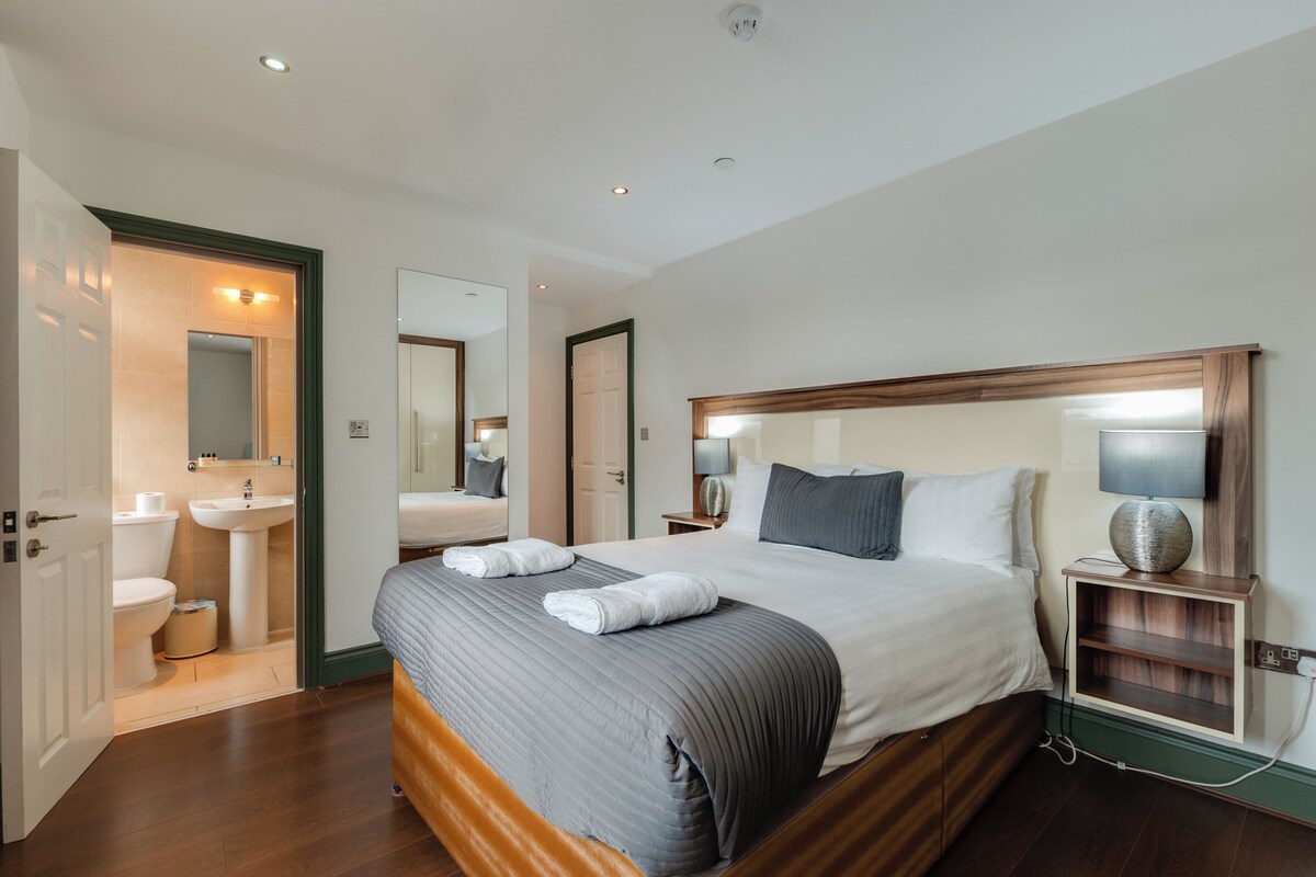 A hotel room with a large bed, bedside table, lamp, and a view of the bathroom.