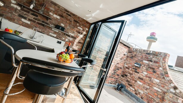A modern kitchen with a brick wall, a dining area with a table and chairs, and a large window offering a view of the city. - Base Serviced Apartments - Sir Thomas Street (Liverpool)