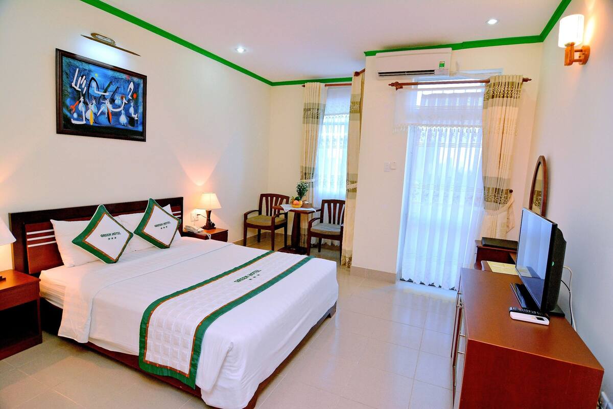 A hotel room with a bed, a desk with a laptop, a chair, a TV, and a painting on the wall.
