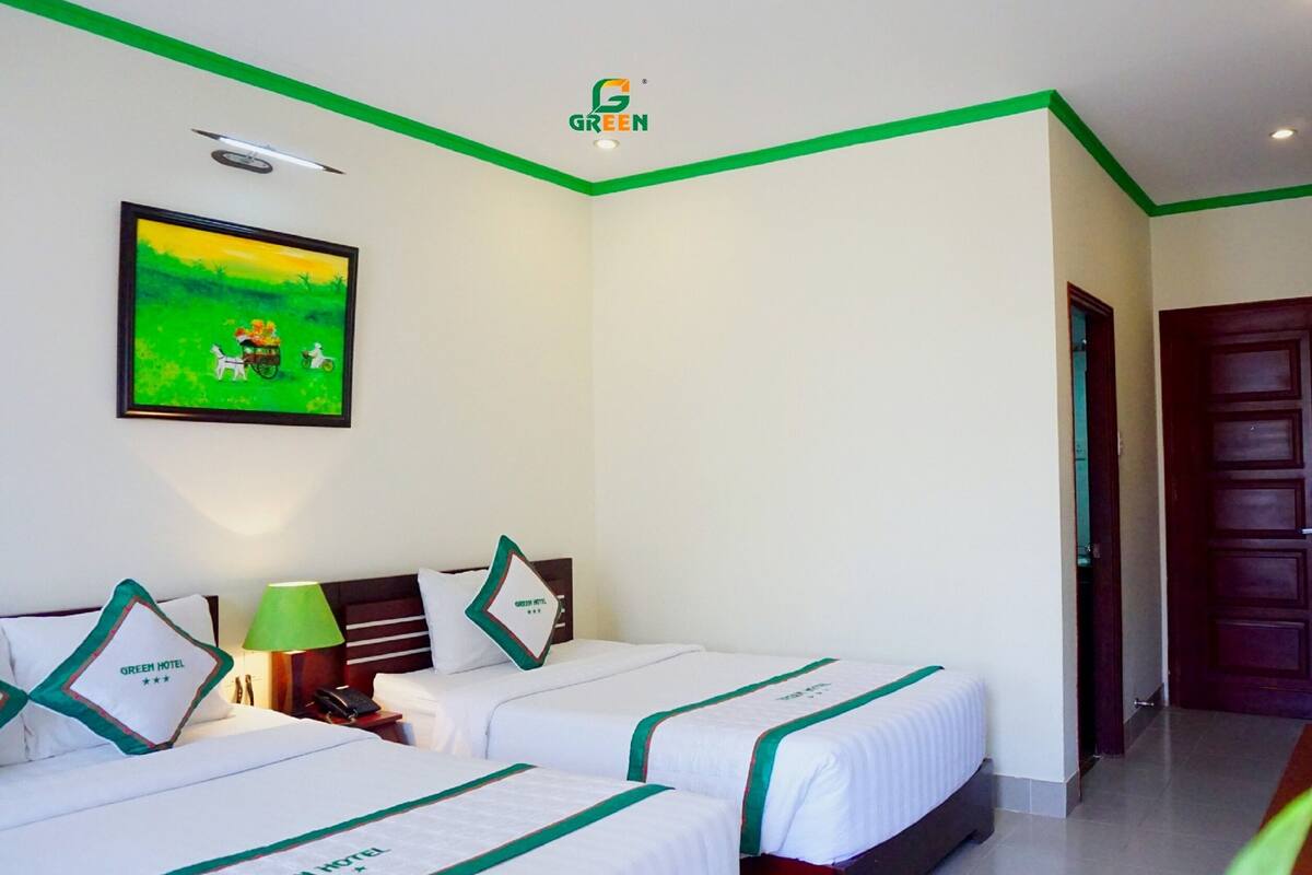 A hotel room with two beds, green accents, a painting of a green landscape, and a door to the right.