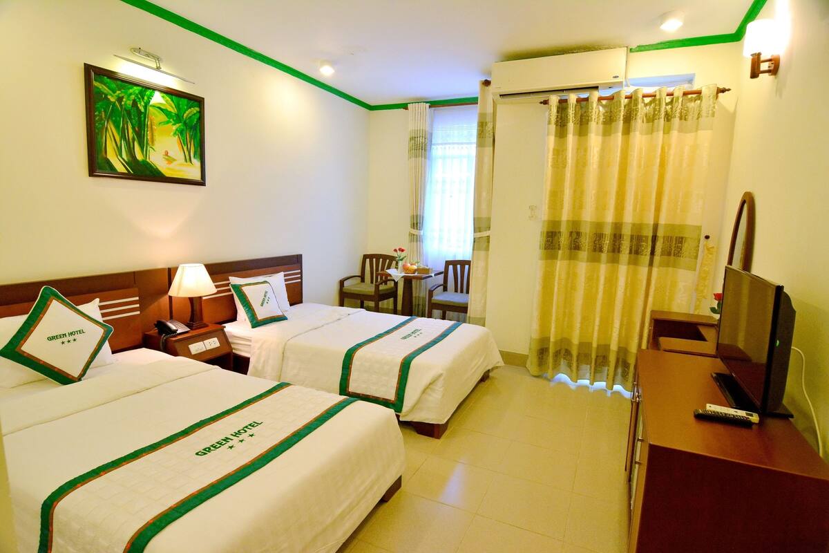 A hotel room with two beds, a desk with a TV, a chair, a table with a vase, and a painting on the wall.