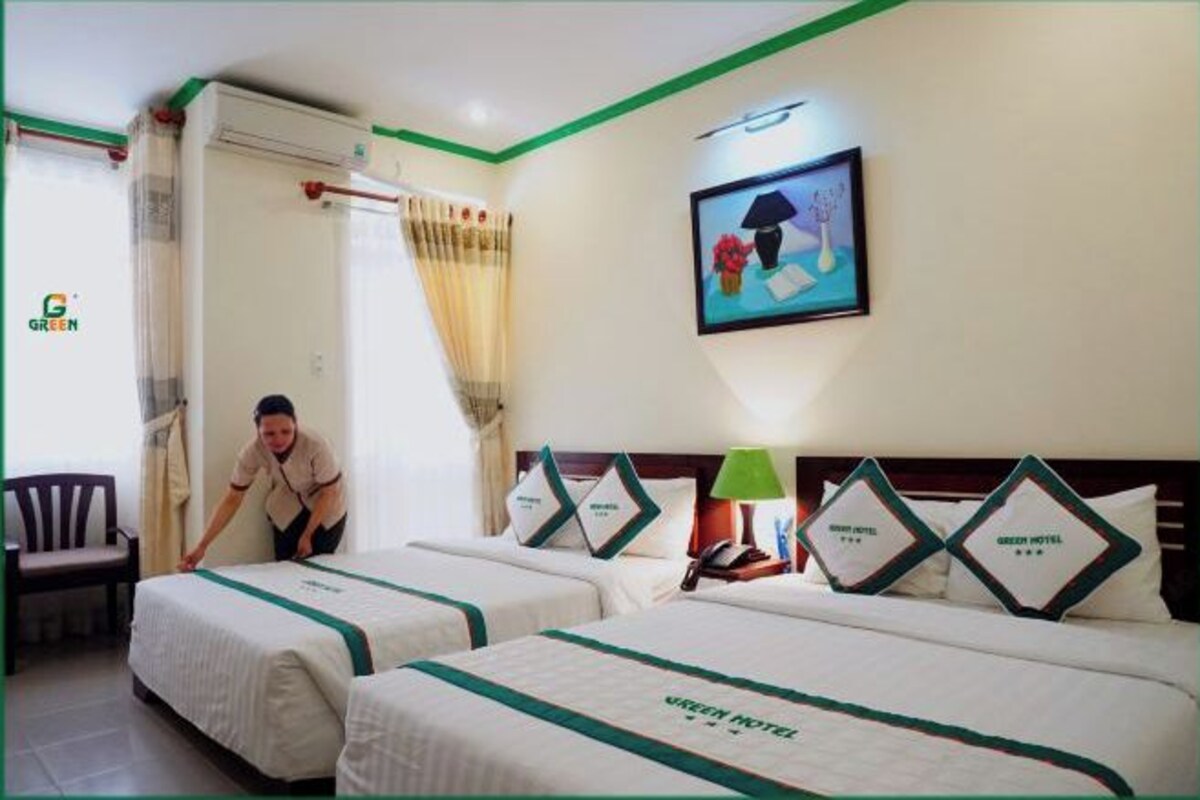 A hotel room with two beds, a green-striped bedspread, a painting on the wall, and a person cleaning.