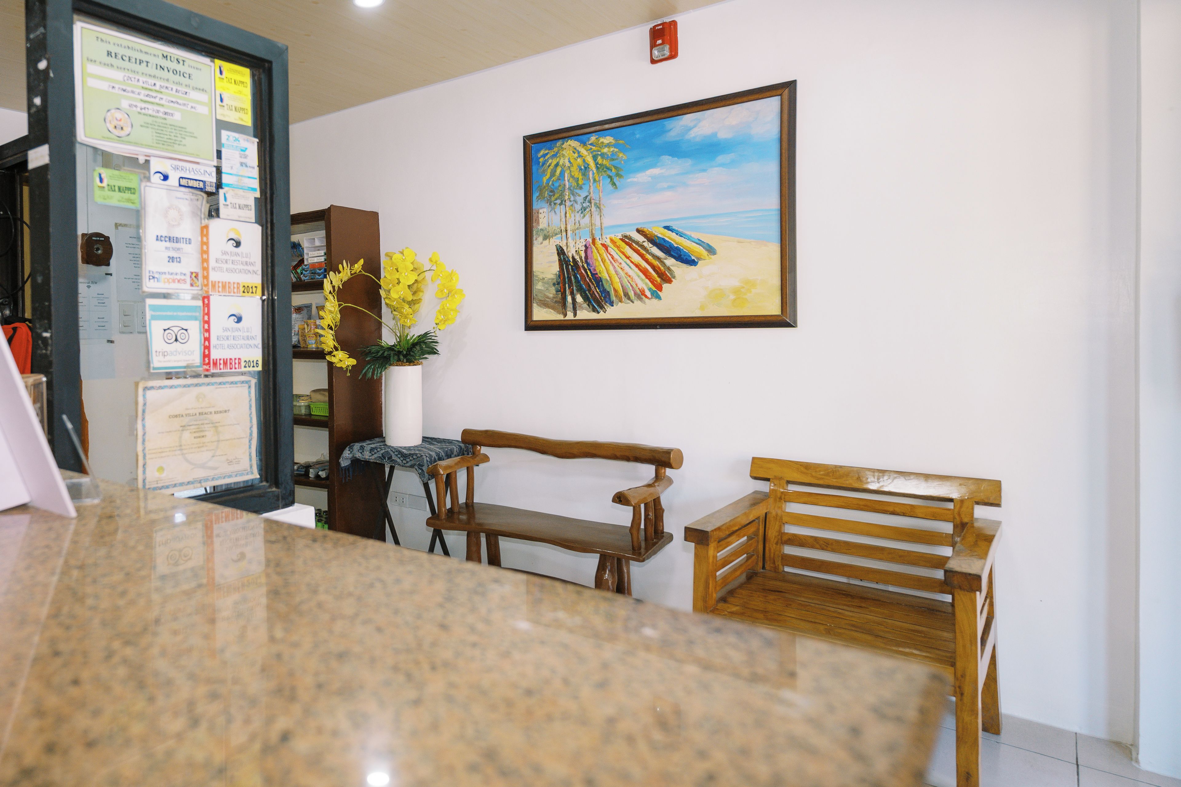 A reception area with a marble counter, wooden benches, a framed painting of a beach scene, and various notices on the wall.