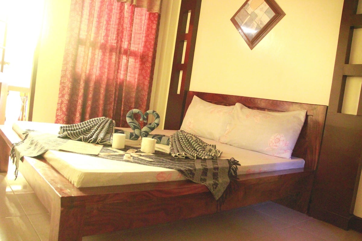 Uma cama bem arrumada com cabeceira de madeira, uma mesa de cabeceira com abajur e uma janela com cortinas vermelhas.