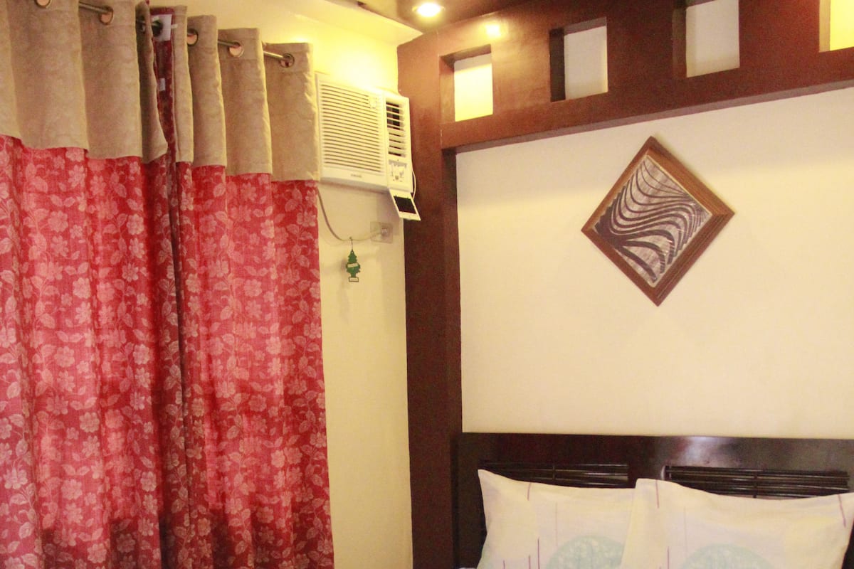 Uma cama com colcha estampada, cabeceira com painel decorativo e janela com cortinas.
