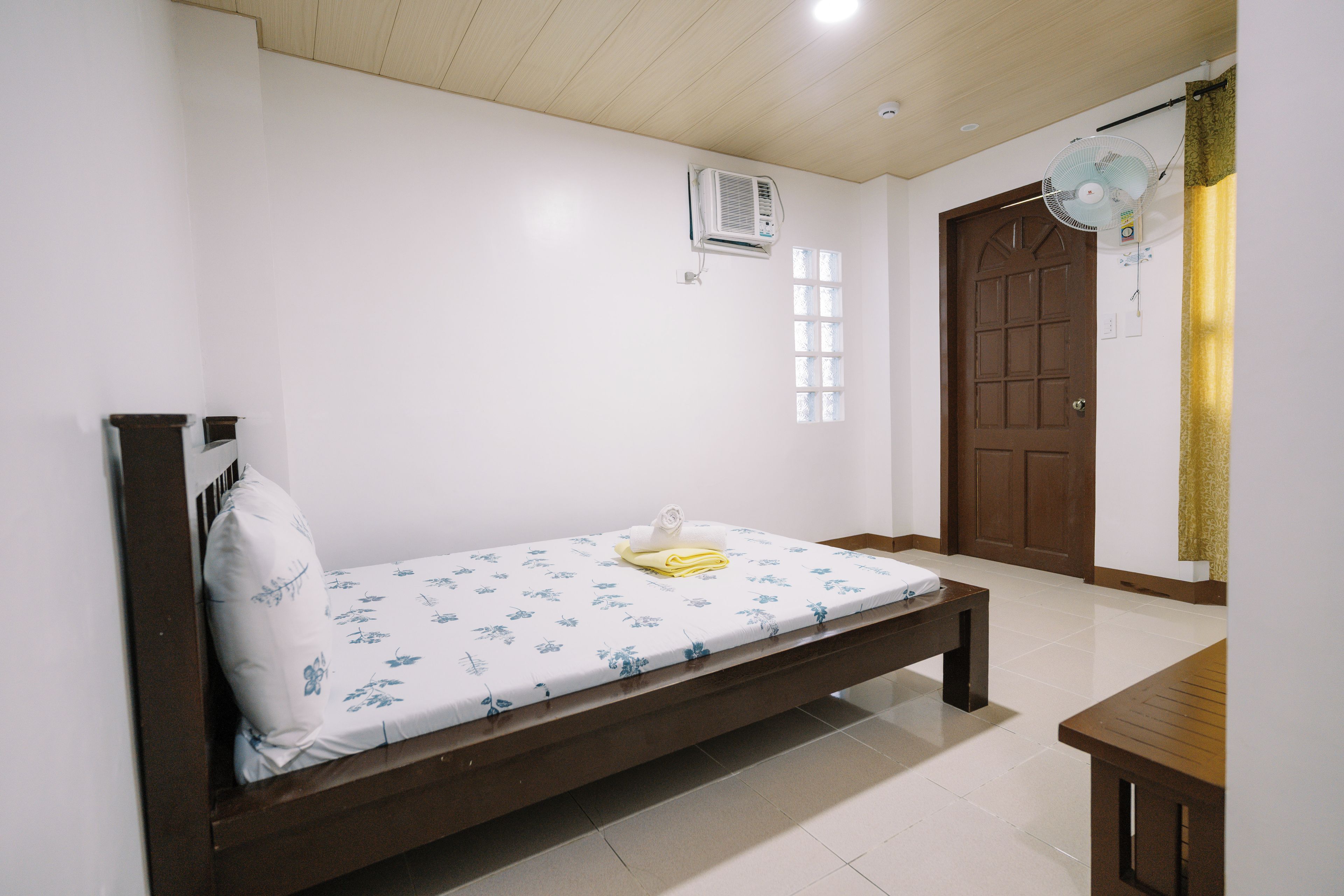 A single-bed room with a wooden frame, a white mattress with blue floral patterns, a bedside table, and a window with a curtain.