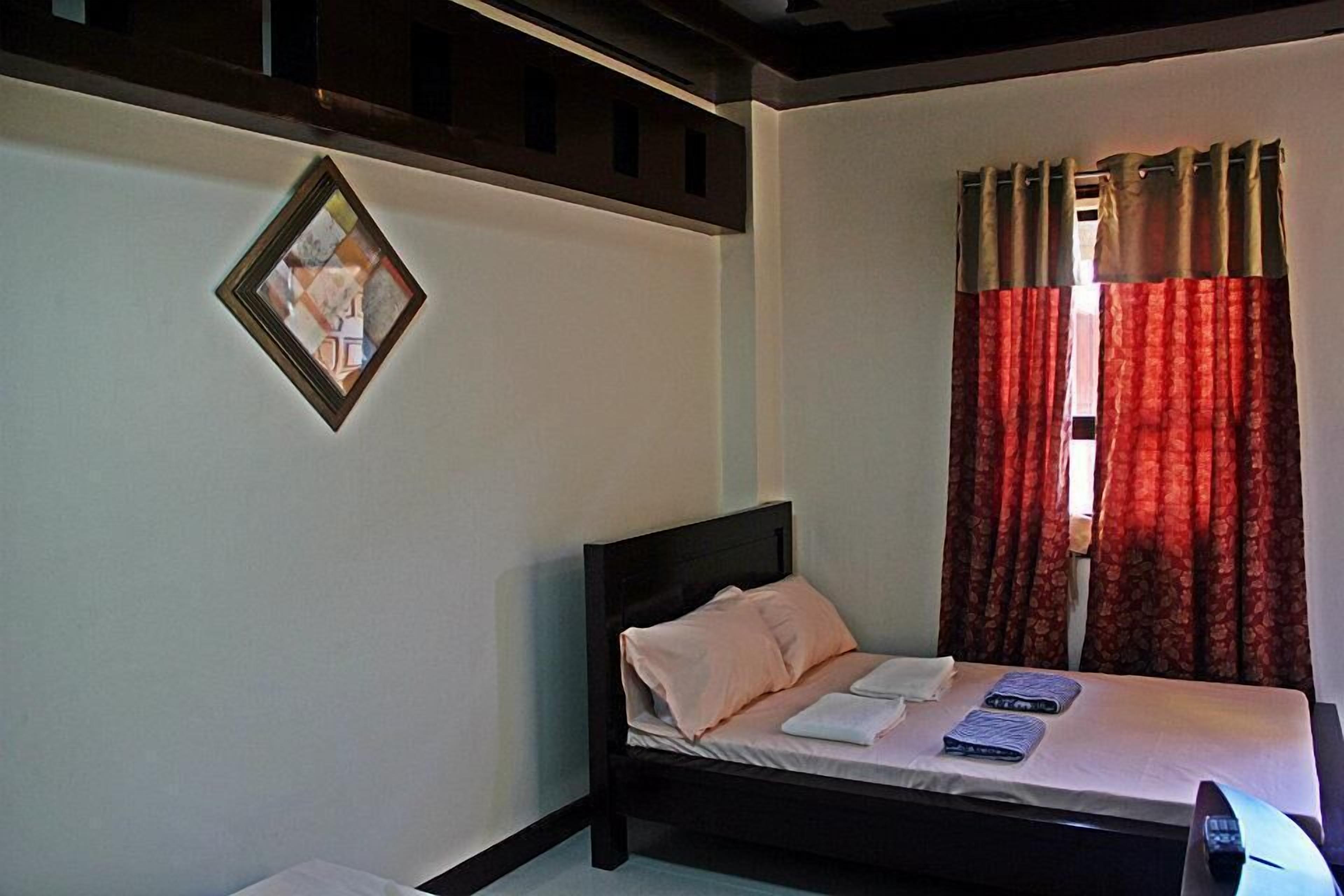A single bed with a headboard, a bedside table, and a window with red curtains.