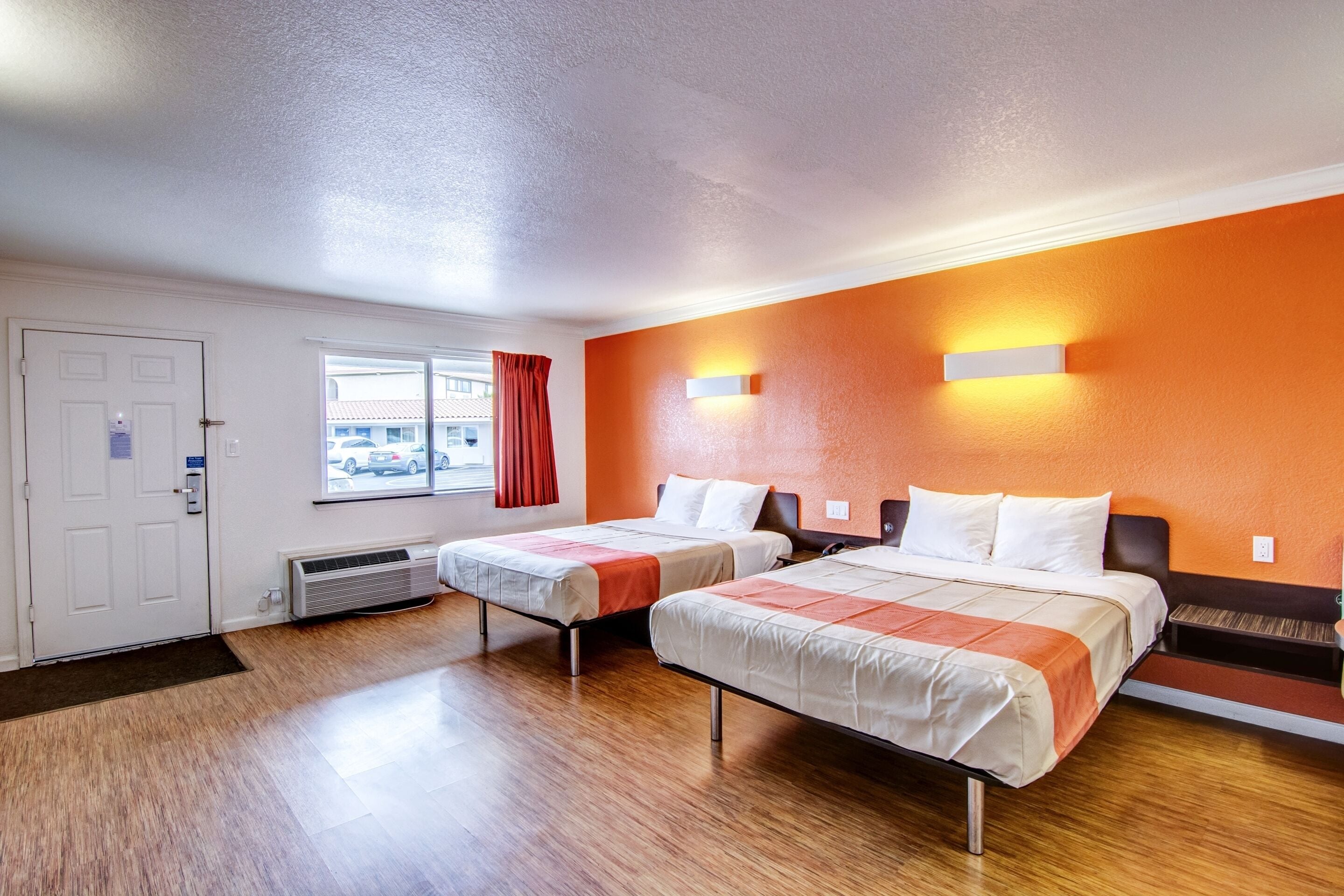 A hotel room with two beds, a window with a view of parked cars, and a door with a handle.