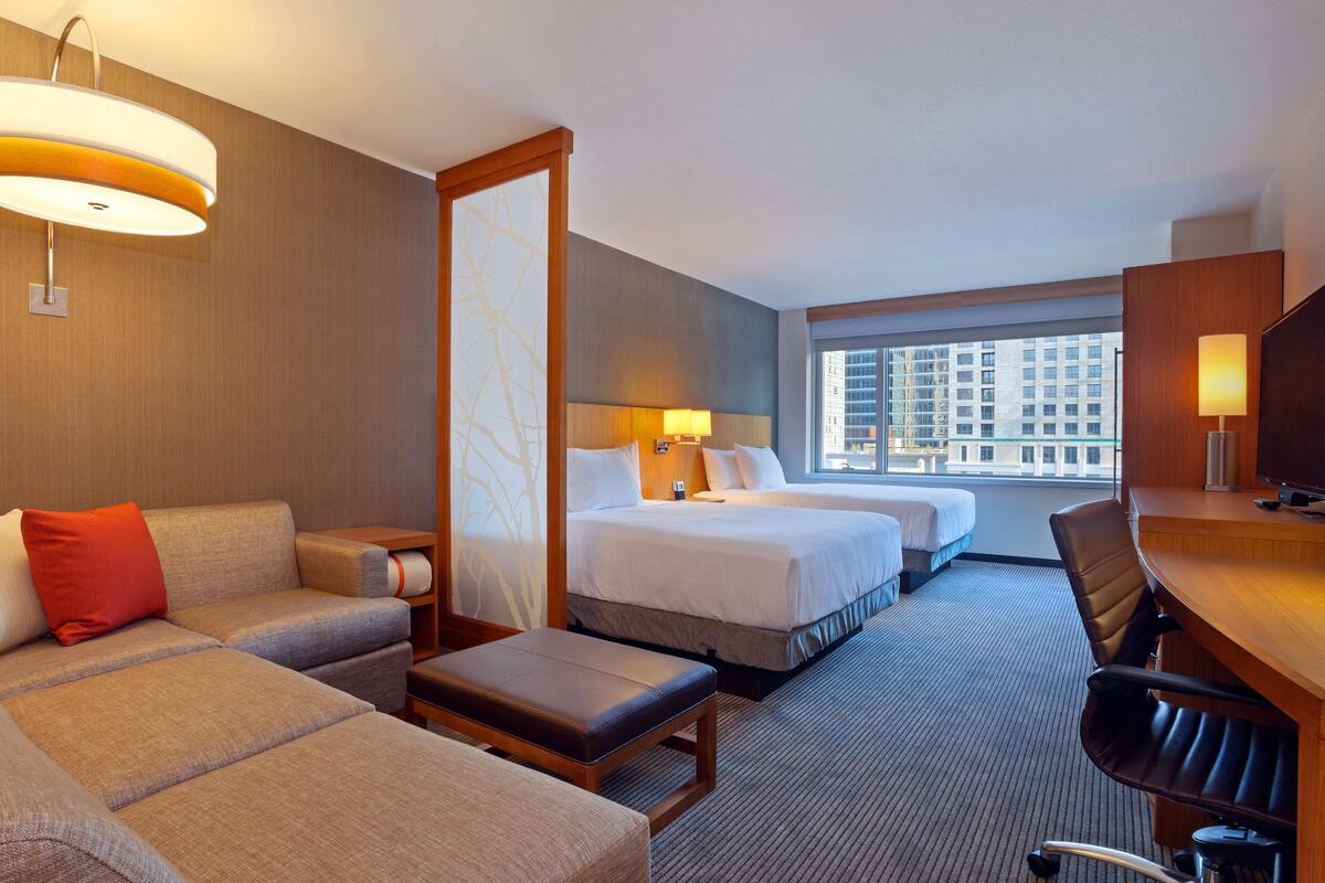 A hotel room with a sofa, two beds, a desk, and a view of a cityscape.