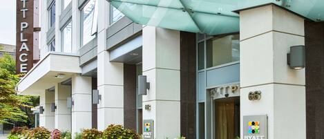 A modern hotel entrance with a glass canopy, a sign reading "HYATT PLACE", and a row of planters with greenery.