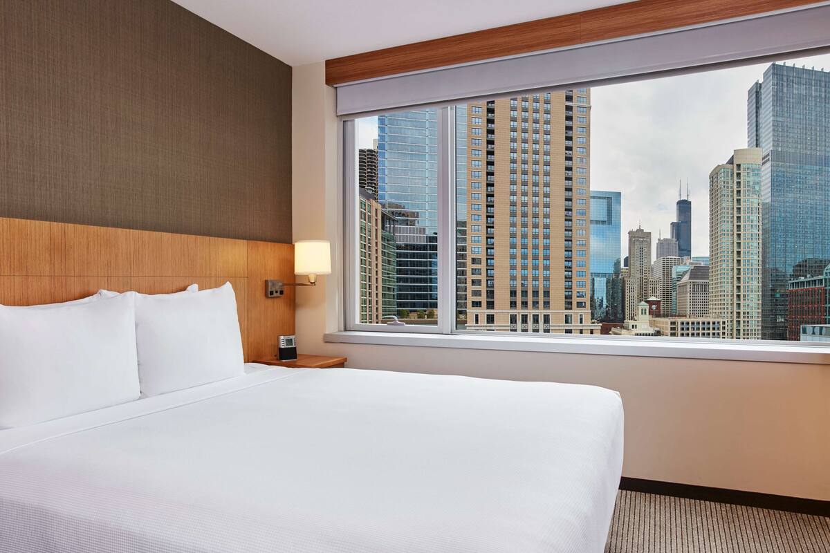 A hotel room with a large bed, a wooden headboard, a nightstand with a lamp, and a window offering a city view.