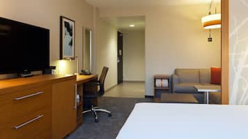 Quarto de hotel com cama, escrivaninha com cadeira, televisão, sofá e luminária.