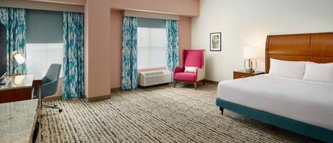 A hotel room with a bed, a desk, a chair, a window with curtains, and a wall-mounted air conditioner.