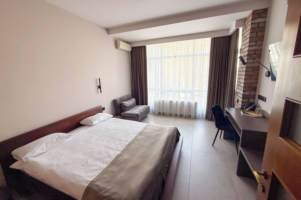 Una habitación de hotel con una cama grande, un escritorio con silla, televisión y ventana con cortinas.