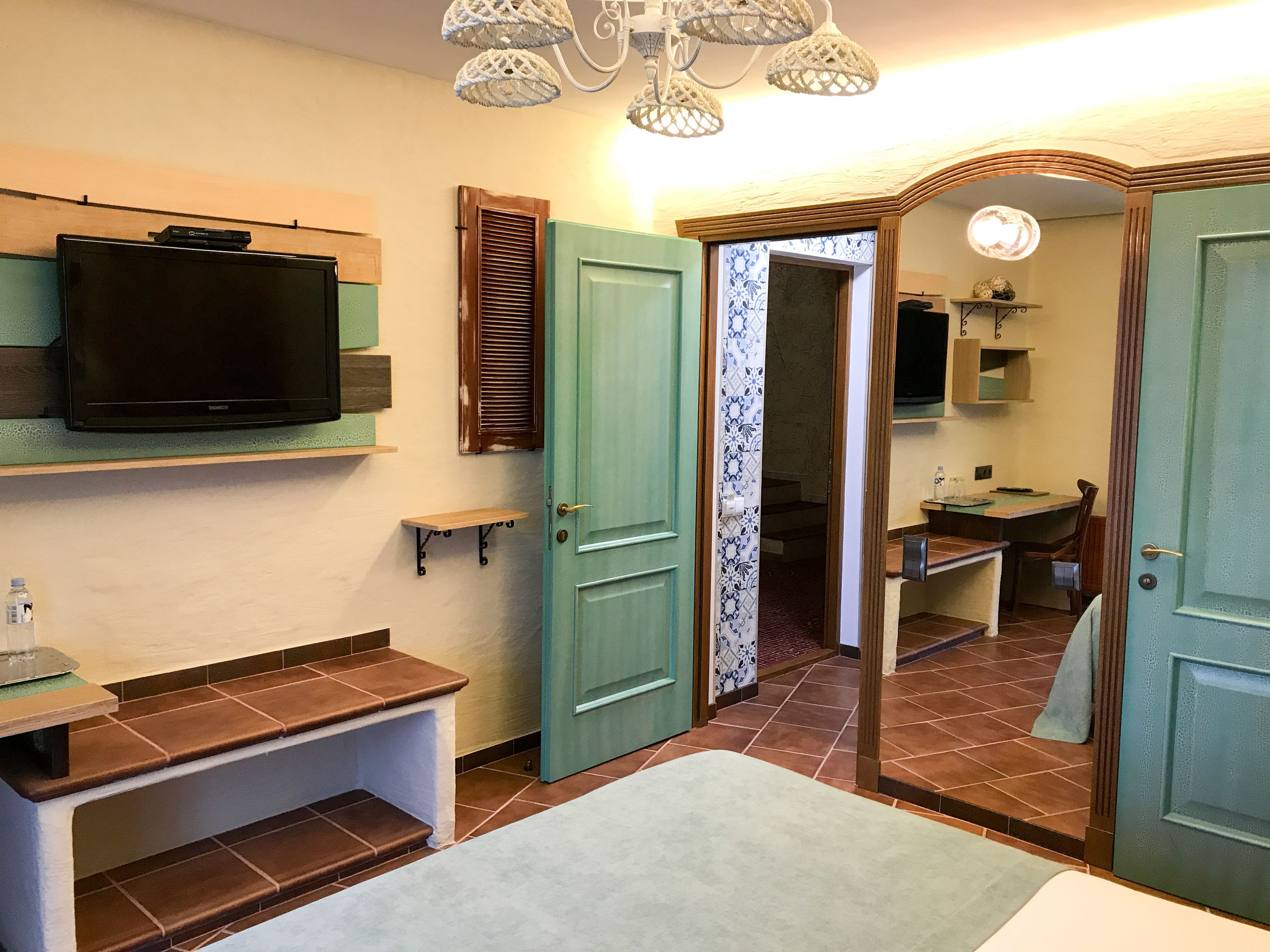 Une chambre d’hôtel équipée d’une télévision, d’une petite étagère, d’un banc, d’une porte, d’une salle de bain avec un lavabo et un miroir, et d’un rideau à motifs.
