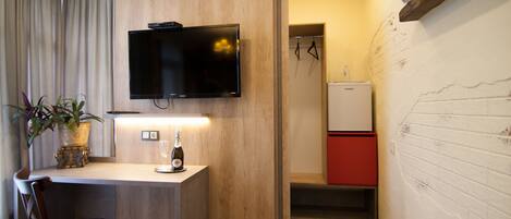 A hotel room with a wooden desk, a television mounted above it, a small table with a bottle and glass, a chair, and a red-framed mirror.