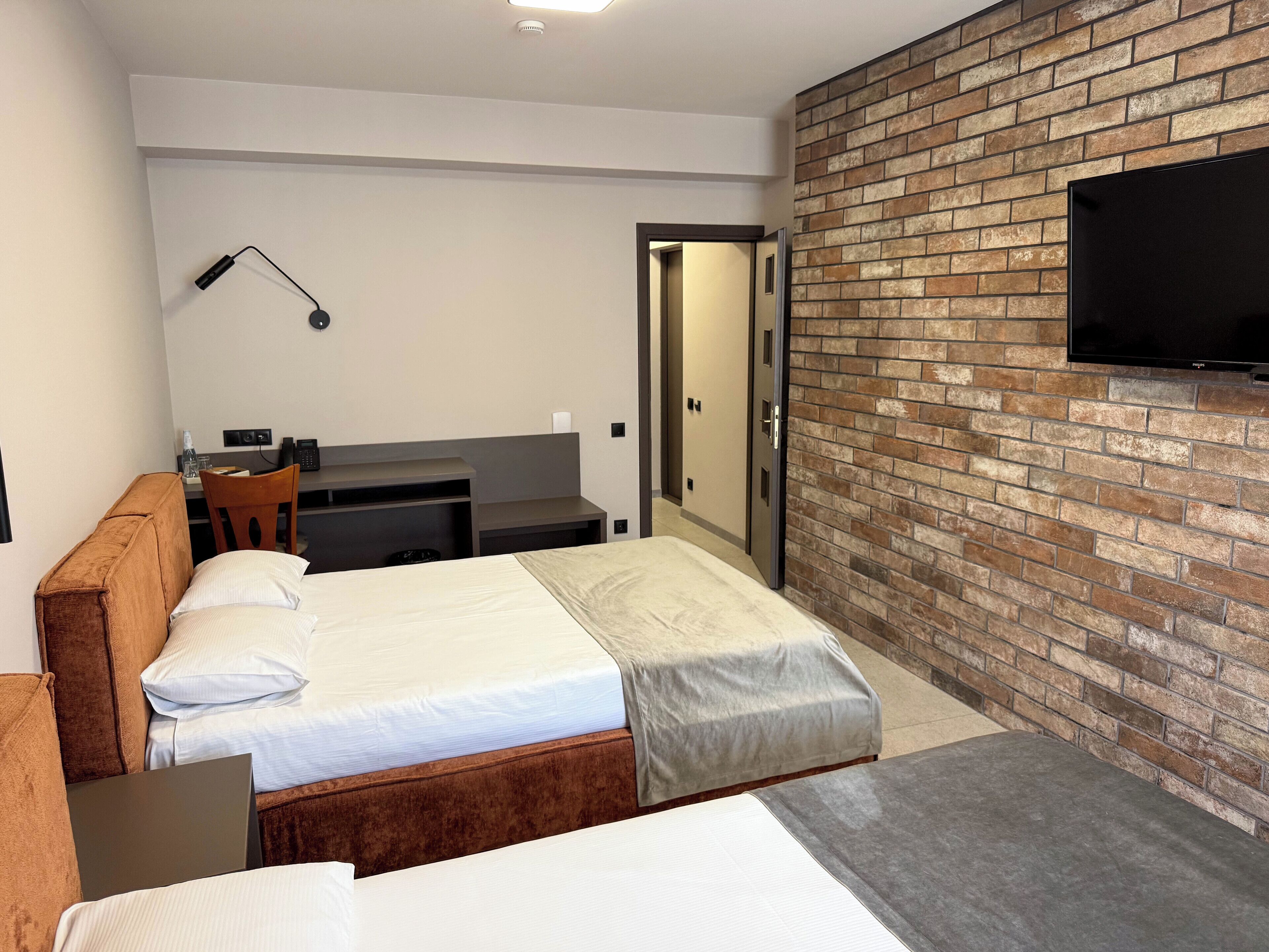 Ein modernes Hotelzimmer mit einer Ziegelfassade, einem flachbildfernseher über einem Schreibtisch und einem separaten Schlafbereich mit Bett und Schreibtisch.