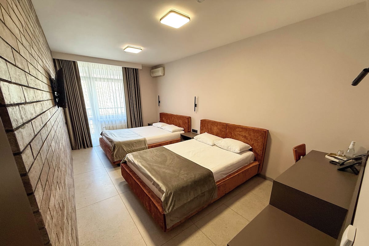 Habitación de hotel con dos camas, un escritorio, una silla, un ventanal con cortinas y un aire acondicionado de pared.