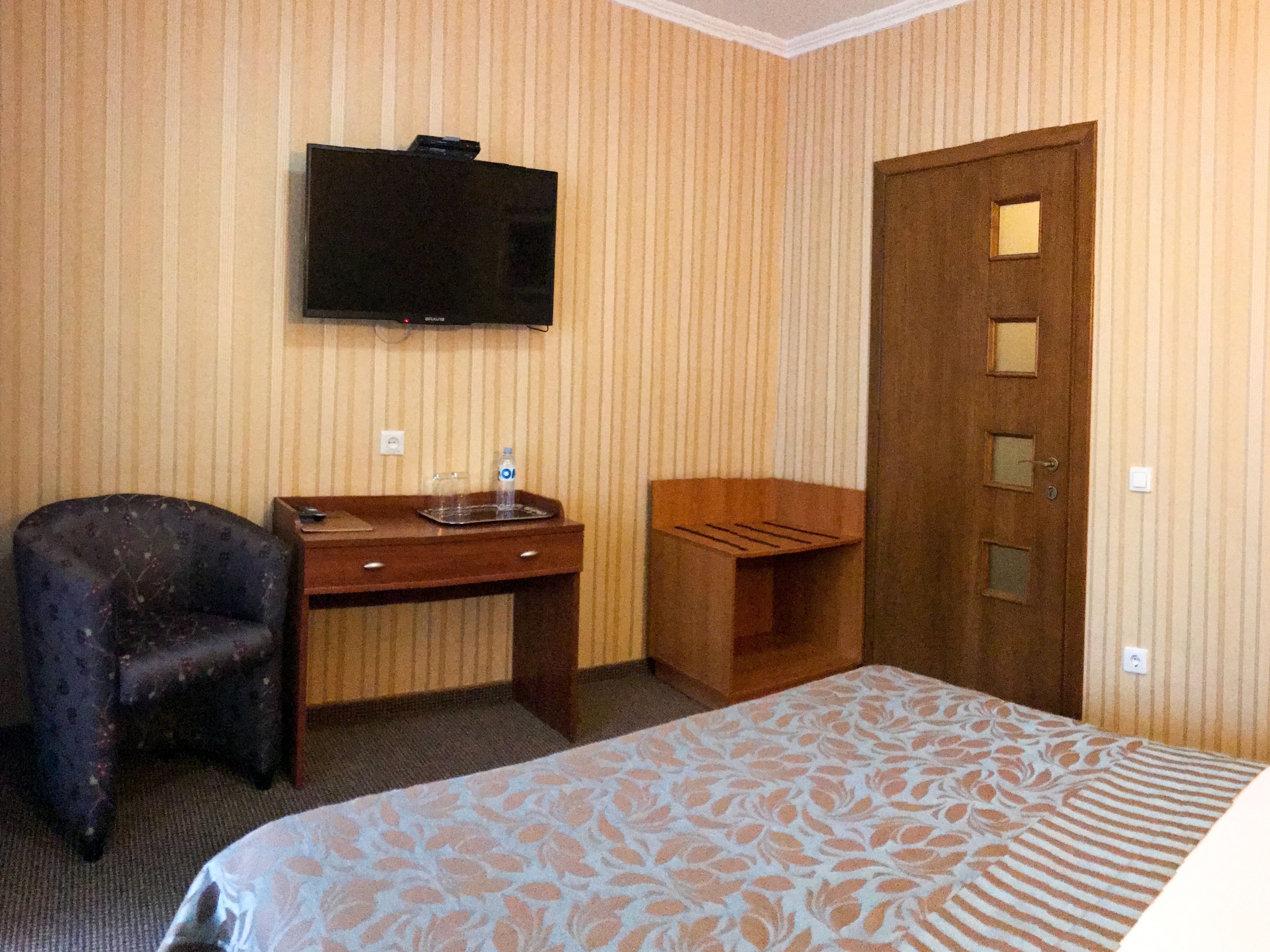 A hotel room with a bed, a desk with a chair, a television mounted on the wall, and a wooden door.