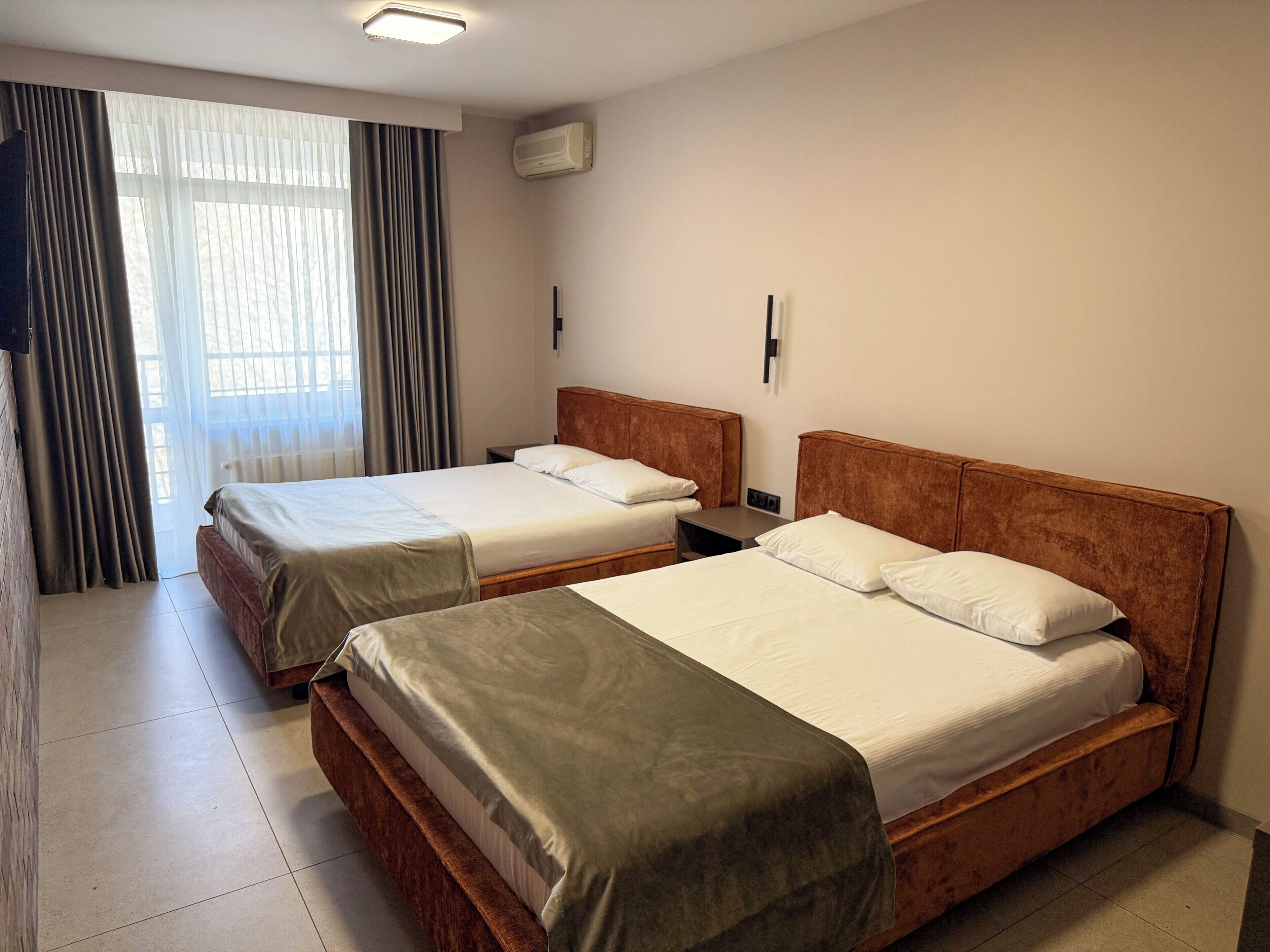 Una camera d'albergo con due letti, una finestra con tende, un condizionatore d'aria e un pavimento piastrellato.