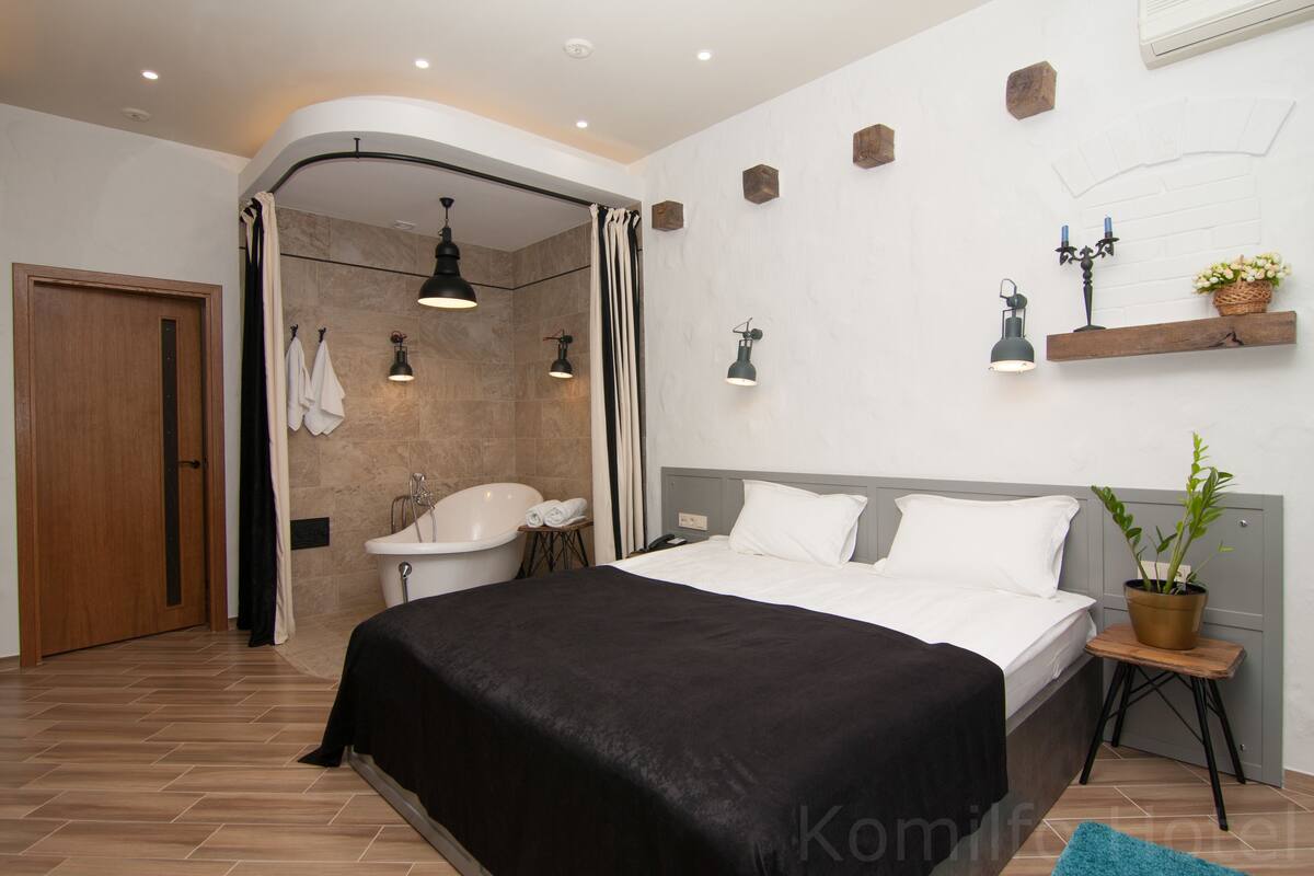 Habitación de hotel moderna con una cama grande, zona de ducha, lavamanos y una pequeña mesa con una planta.