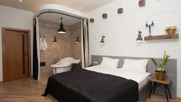 Habitación de hotel moderna con una cama grande, zona de ducha, lavamanos y una pequeña mesa con una planta.