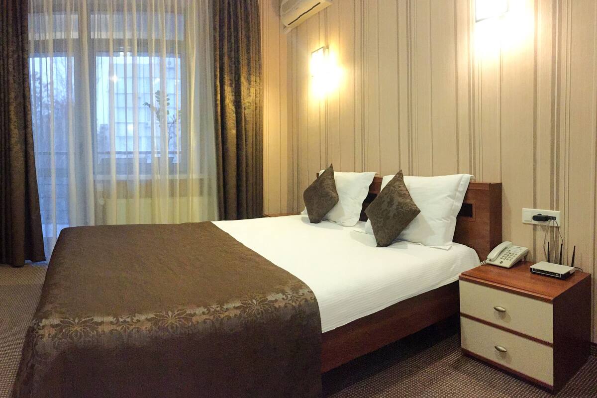 Una habitación de hotel con una cama grande, una mesita de noche con teléfono y lámpara, y una ventana con cortinas.