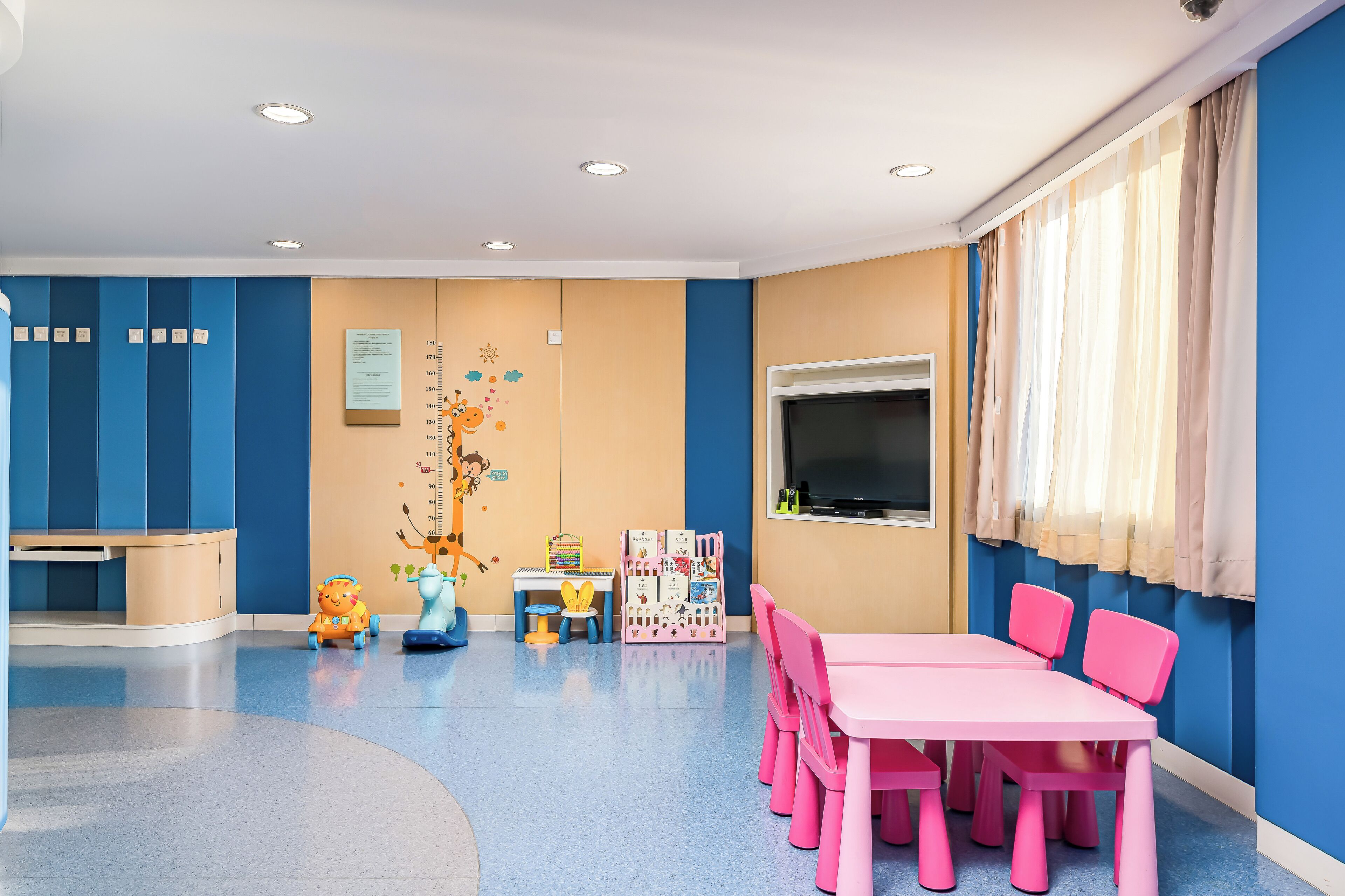 Ein Kinderspielzimmer mit blauen und beigen Farbtönen, ausgestattet mit einem rosafarbenen Tisch und Stühlen, einem Spielbereich mit Spielzeug und einer Größenangabe an der Wand.