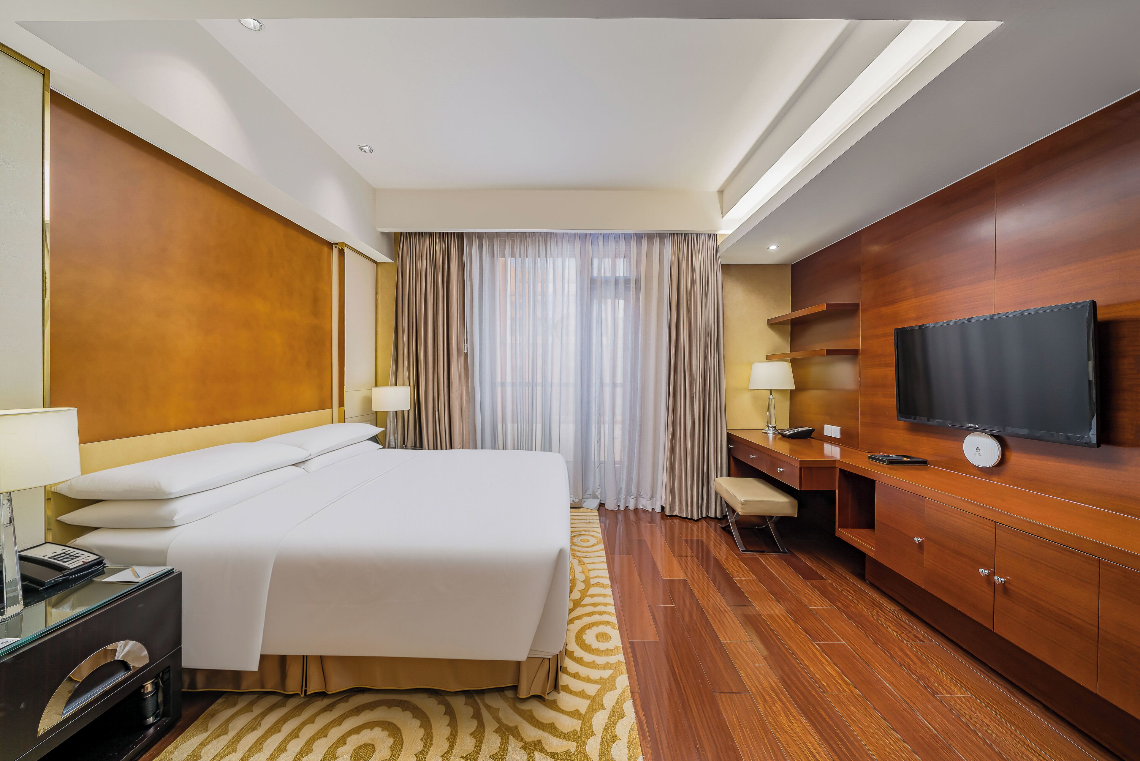 A hotel room with a bed, bedside table, lamp, TV, wooden cabinet, and a large window with curtains.