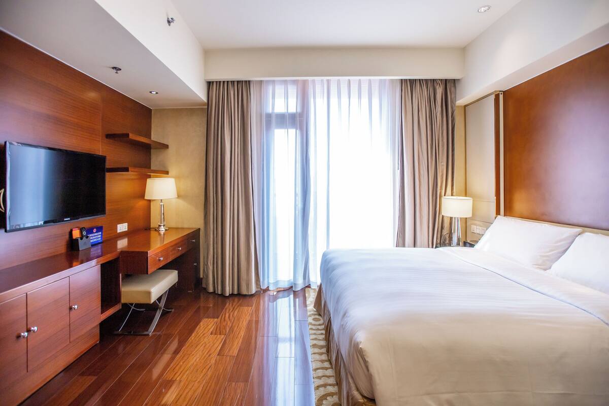 A hotel room with a bed, a TV, a desk, a chair, and a window with curtains.