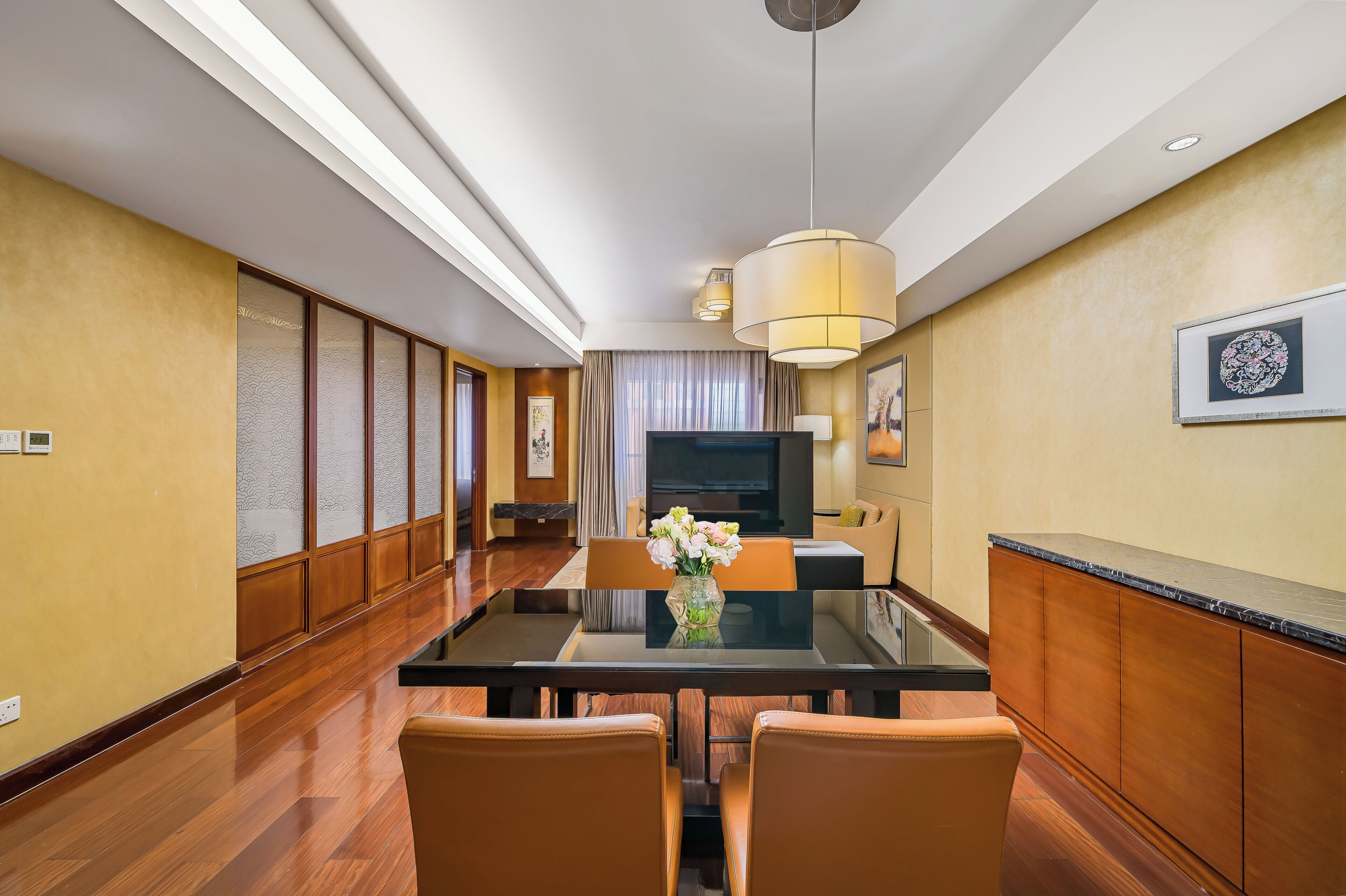 Habitación de hotel con zona de comedor que incluye una mesa de vidrio, sillas de cuero, un jarrón con flores, un televisor de pantalla plana y un ventanal con cortinas.