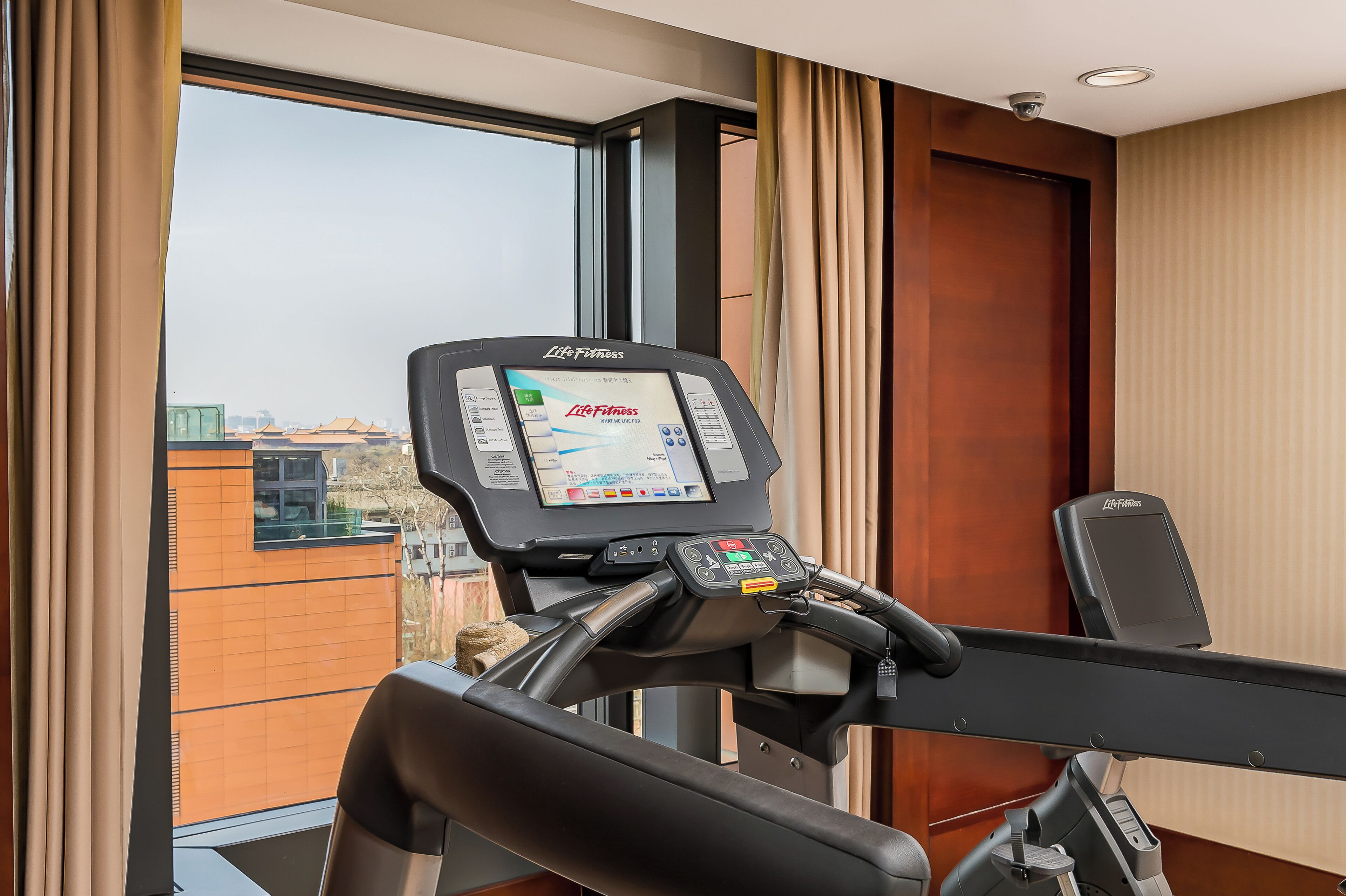 A treadmill with a digital display, a window with curtains, and a view of buildings outside.