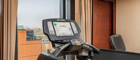 A treadmill with a digital display, a window with curtains, and a view of buildings outside.