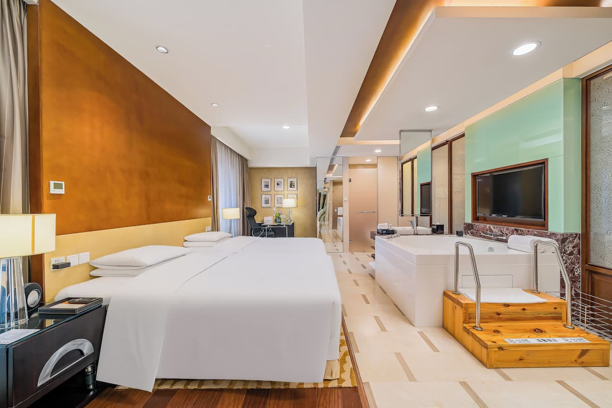 A hotel room with a large bed, a desk with a lamp, a TV, a bathroom with a bathtub, and a shower area.